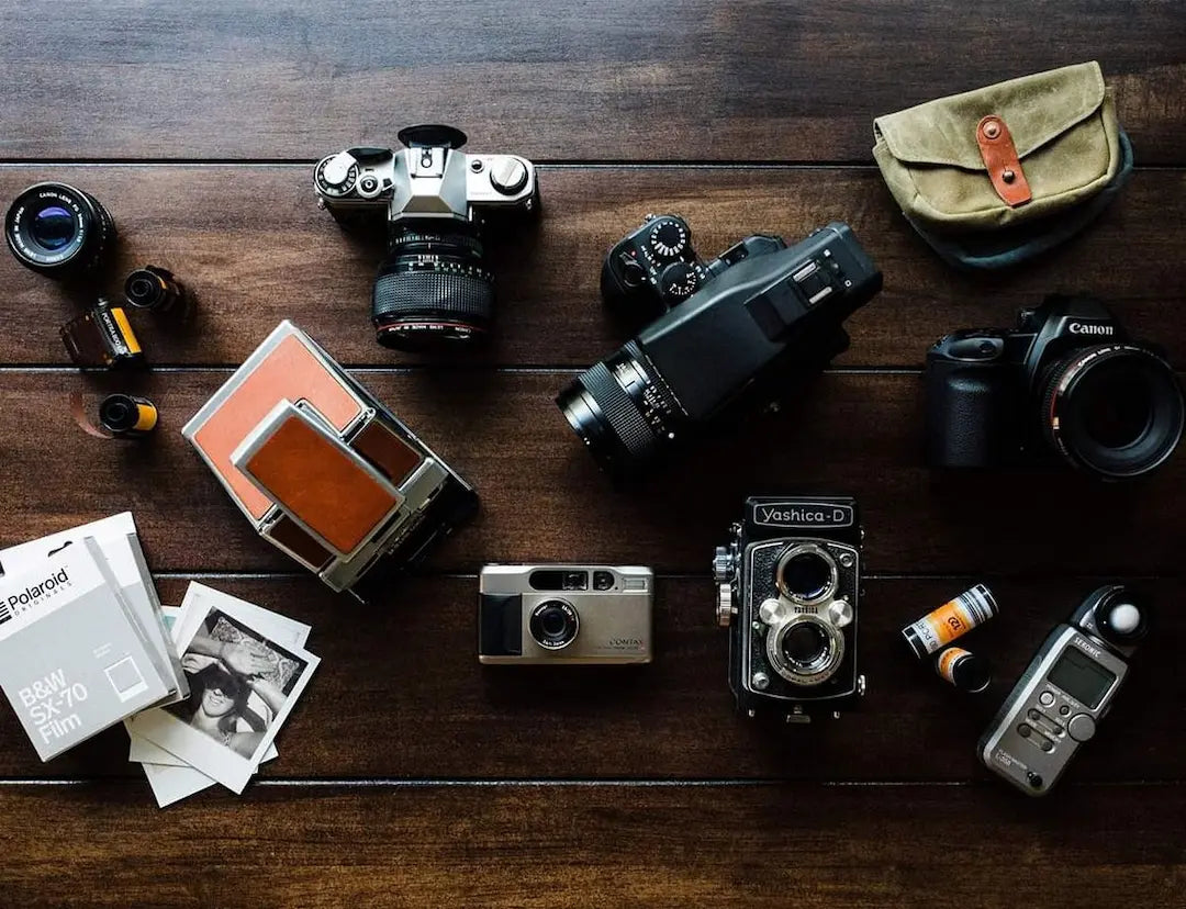 Vue de dessus d'une collection d'appareils photo argentiques incluant un Yashica-D, un Contax T2, un Polaroid SX-70 et des pellicules 35mm sur une table en bois.
